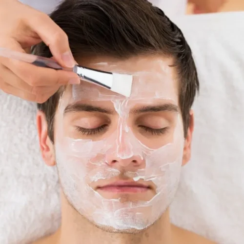 A man receiving a facial treatment with a white cream mask being applied using a brush, highlighting the use of essential skincare products for healthy and refreshed skin.