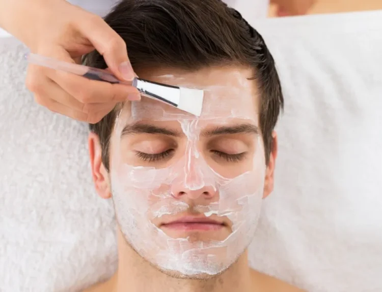 A man receiving a facial treatment with a white cream mask being applied using a brush, highlighting the use of essential skincare products for healthy and refreshed skin.