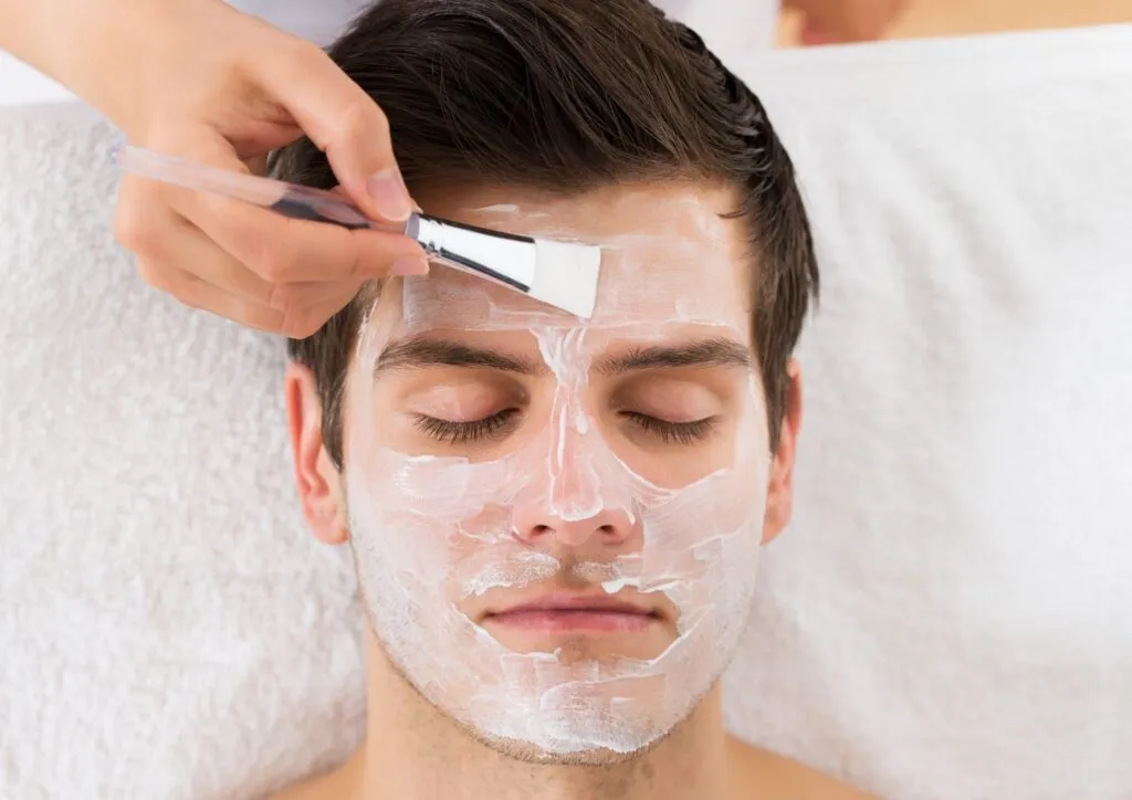 A man receiving a facial treatment with a white cream mask being applied using a brush, highlighting the use of essential skincare products for healthy and refreshed skin.