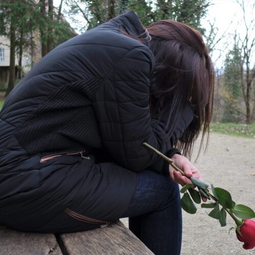 A girl in a black puffy jacket sits on a wooden bench, holding a red rose with a sorrowful demeanor, possibly reflecting on a narcissist mother. Background shows a stone building and leafless trees on a cloudy day.
