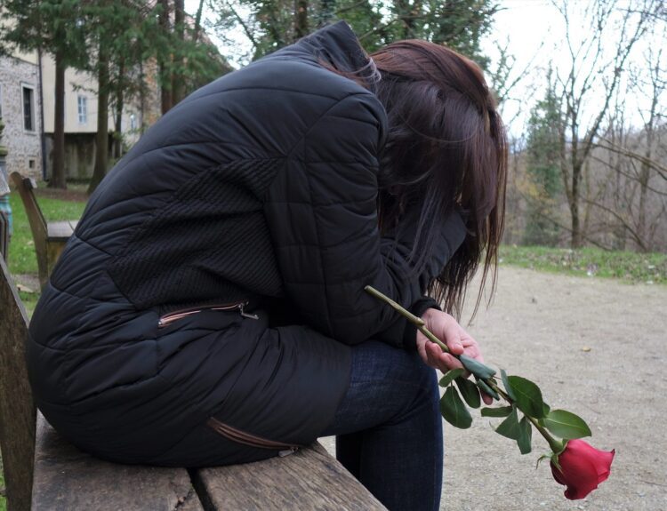 A girl in a black puffy jacket sits on a wooden bench, holding a red rose with a sorrowful demeanor, possibly reflecting on a narcissist mother. Background shows a stone building and leafless trees on a cloudy day.
