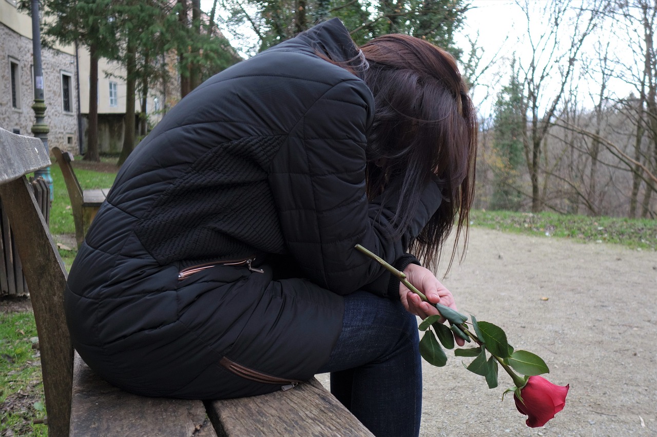 A girl in a black puffy jacket sits on a wooden bench, holding a red rose with a sorrowful demeanor, possibly reflecting on a narcissist mother. Background shows a stone building and leafless trees on a cloudy day.