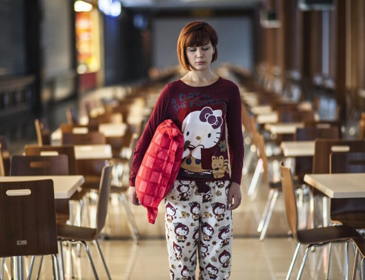 A girl holding a crimson cushion in the center of the cafeteria is sleepwalking in her pajamas.