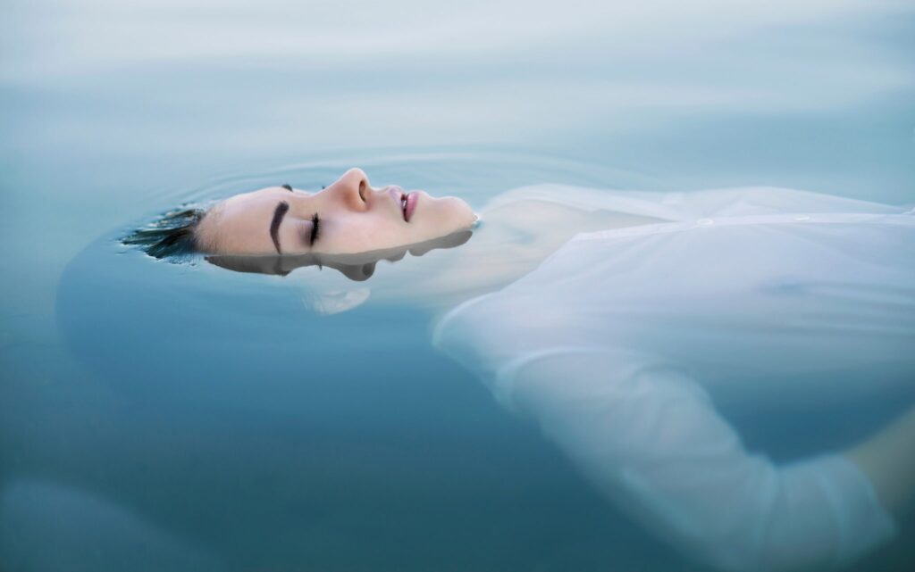 As though floating through a lucid dream, a stunning woman is serenely floating on water, her eyes closed, her mouth above the surface, and her breathing easy.